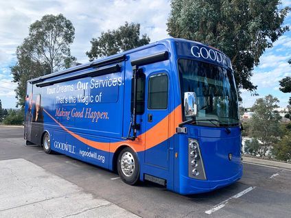 Blue Goodwill bus parked outdoors in a lot, with trees and a partly cloudy sky in the background