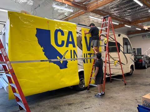 People installing a large yellow and blue truck wrap inside a garage, with a ladder and vehicle nearby