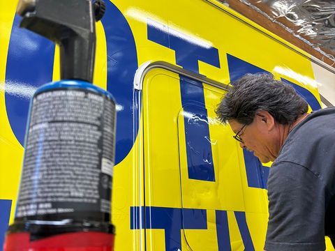 Person painting large blue letters on a bright yellow sign with a spray can