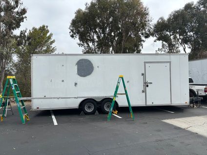 White utility trailer with a door and round vent, flanked by two green ladders in a parking lot.