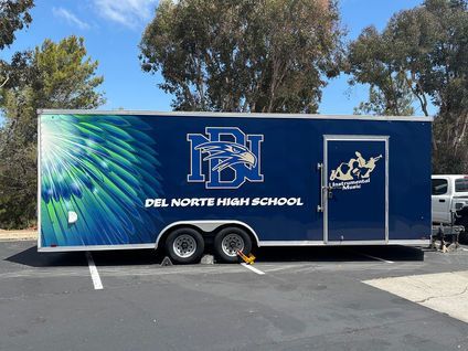 Blue Del Norte High School trailer with school logo parked in a lot under trees and blue sky