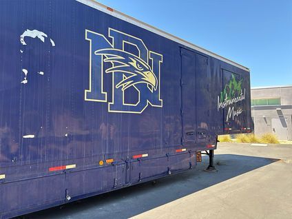 Blue trailer with a large white-and-gold logo parked beside a building on a sunny street.