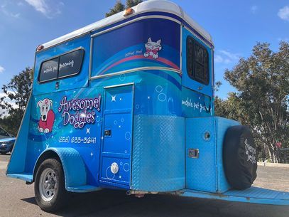 Blue pet grooming trailer with cartoon logos and a rear spare tire parked outdoors under a blue sky