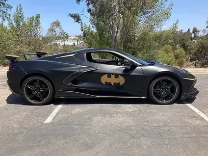 Black sports car with a gold Batman logo on the door, parked on a road near trees.