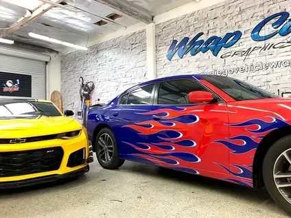 Two customized sports cars in a garage, one yellow and black, one blue with red flames.