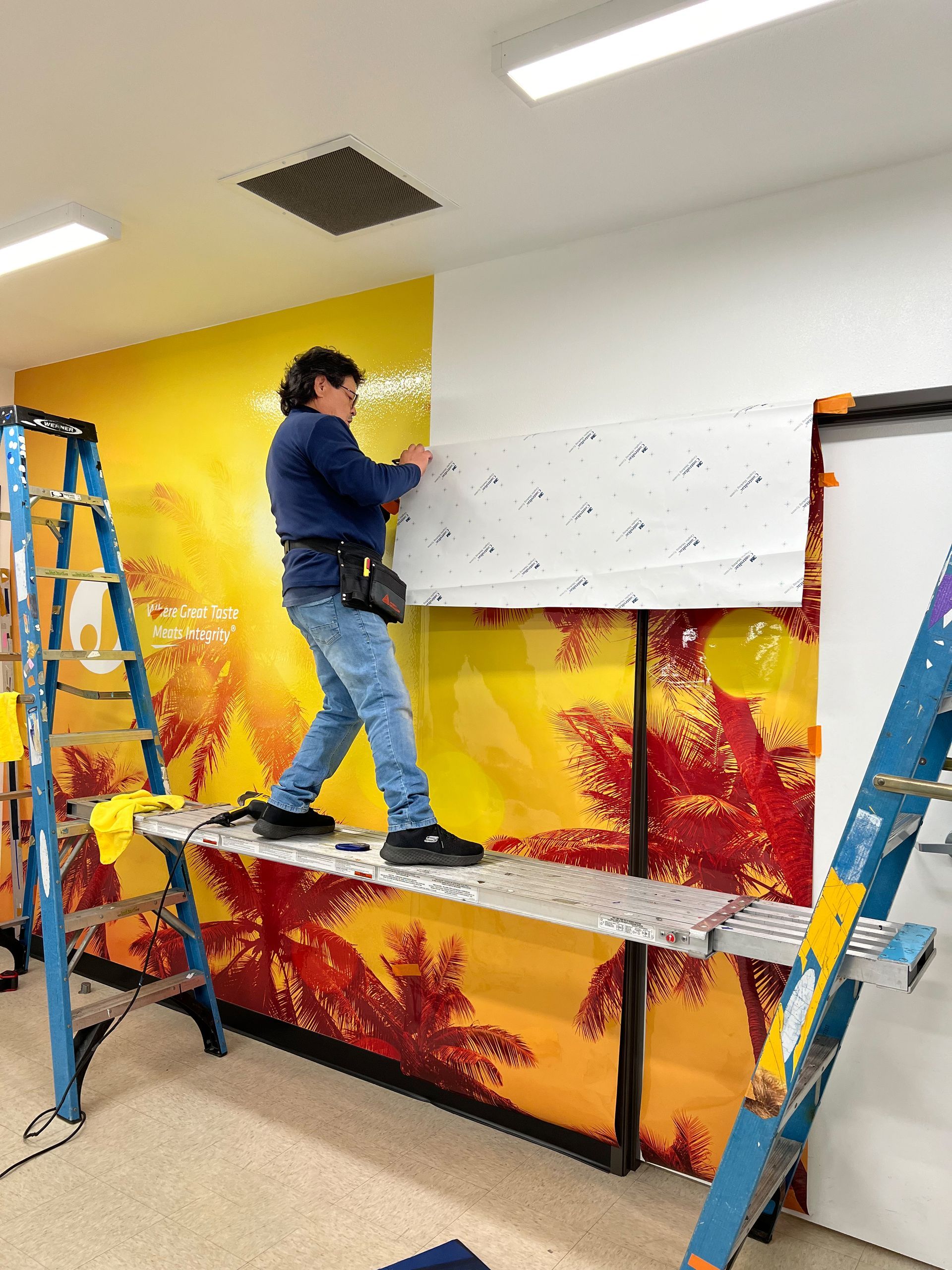 Person hanging white wallpaper on a bright yellow wall, standing on a blue ladder and scaffold.