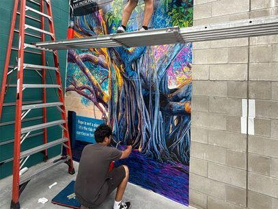 An artist painting a colorful tree mural on a wall beside a ladder in an indoor work area