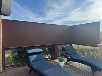 Two black lounge chairs on a wooden deck, overlooking greenery under a large brown privacy screen.