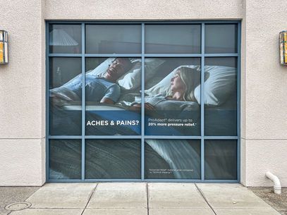 Billboard with sleeping people and the text ACHES & PAINS on a building window panel