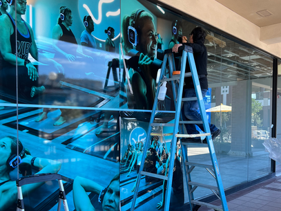 Workers on a ladder installing a large blue mural on a glass storefront wall