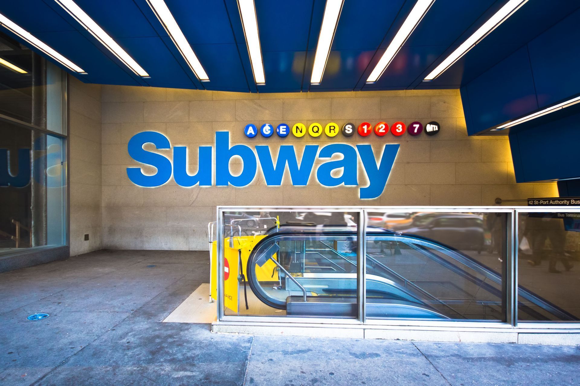 Subway entrance with blue lettering on a beige wall. Escalator visible, blue ceiling, and white lights.