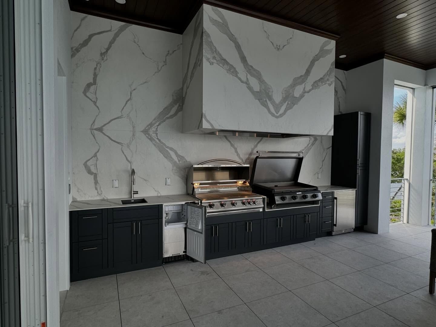 Outdoor patio kitchen with black cabinets, marble-look backsplash, grill, and pizza oven under a roof