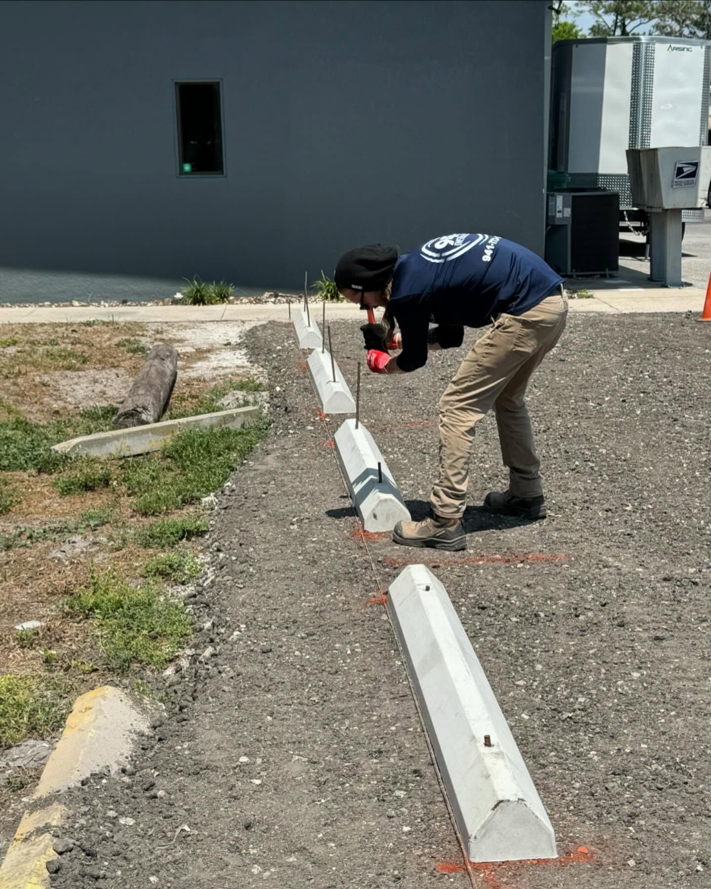 A person kneeling, using a tool to install concrete parking stops in a gravel area.