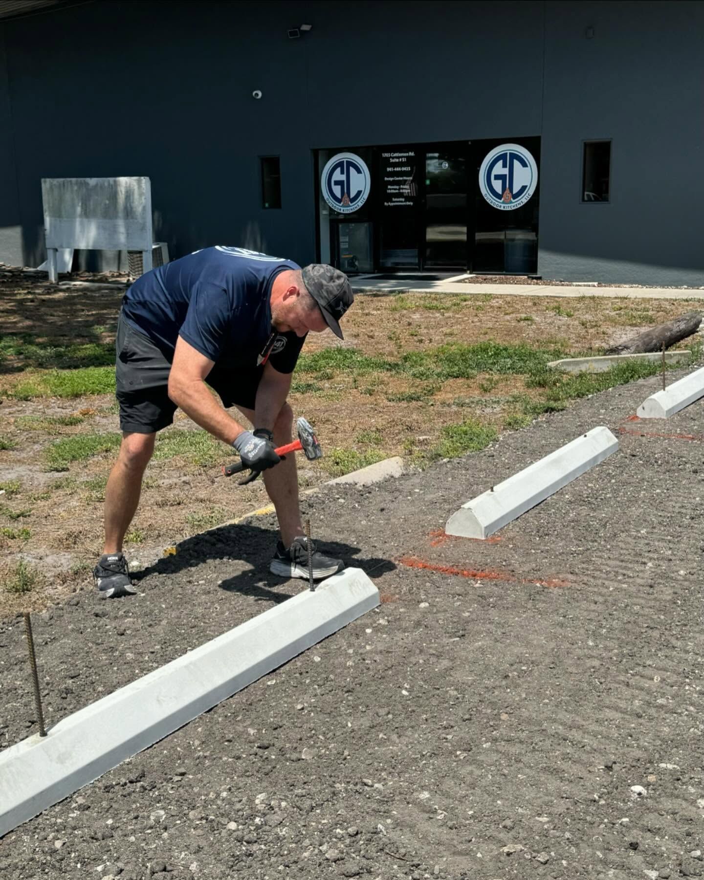 A person hammers a stake into the ground near a parking curb in front of a building with the letters 