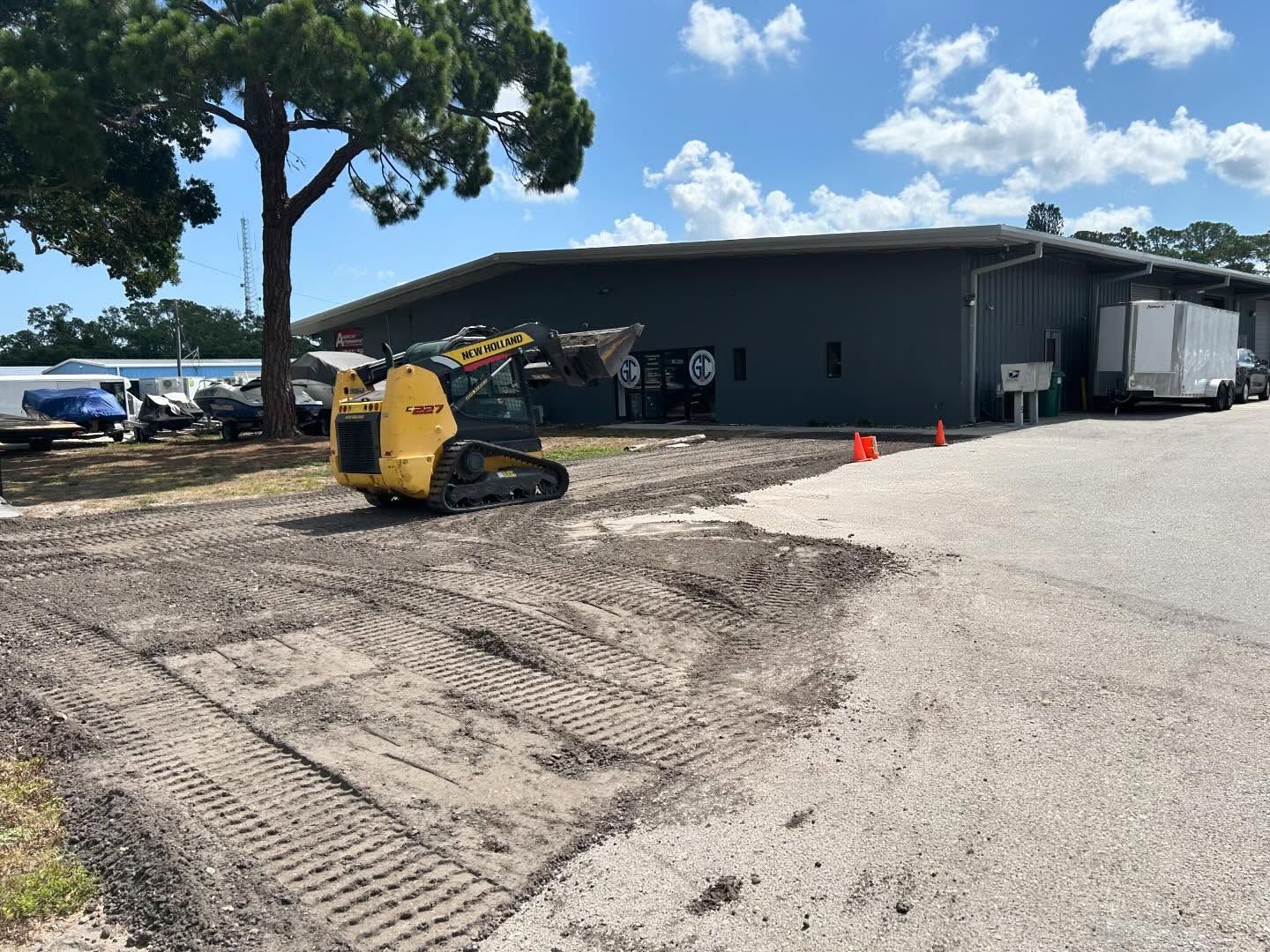 Yellow skid steer on gravel near a dark building and parked vehicles under a partly cloudy sky.