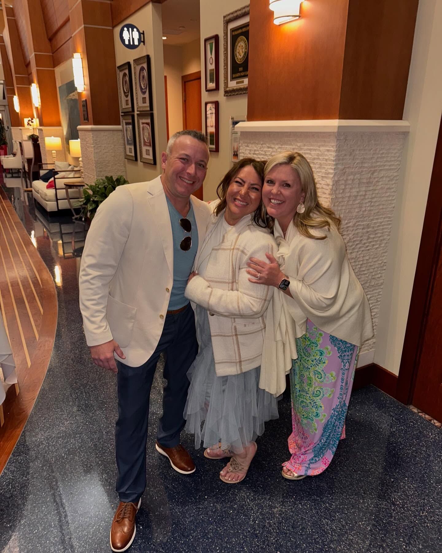 Three people pose indoors: man in cream jacket, women in light blue skirt and colorful shawl, smiling.