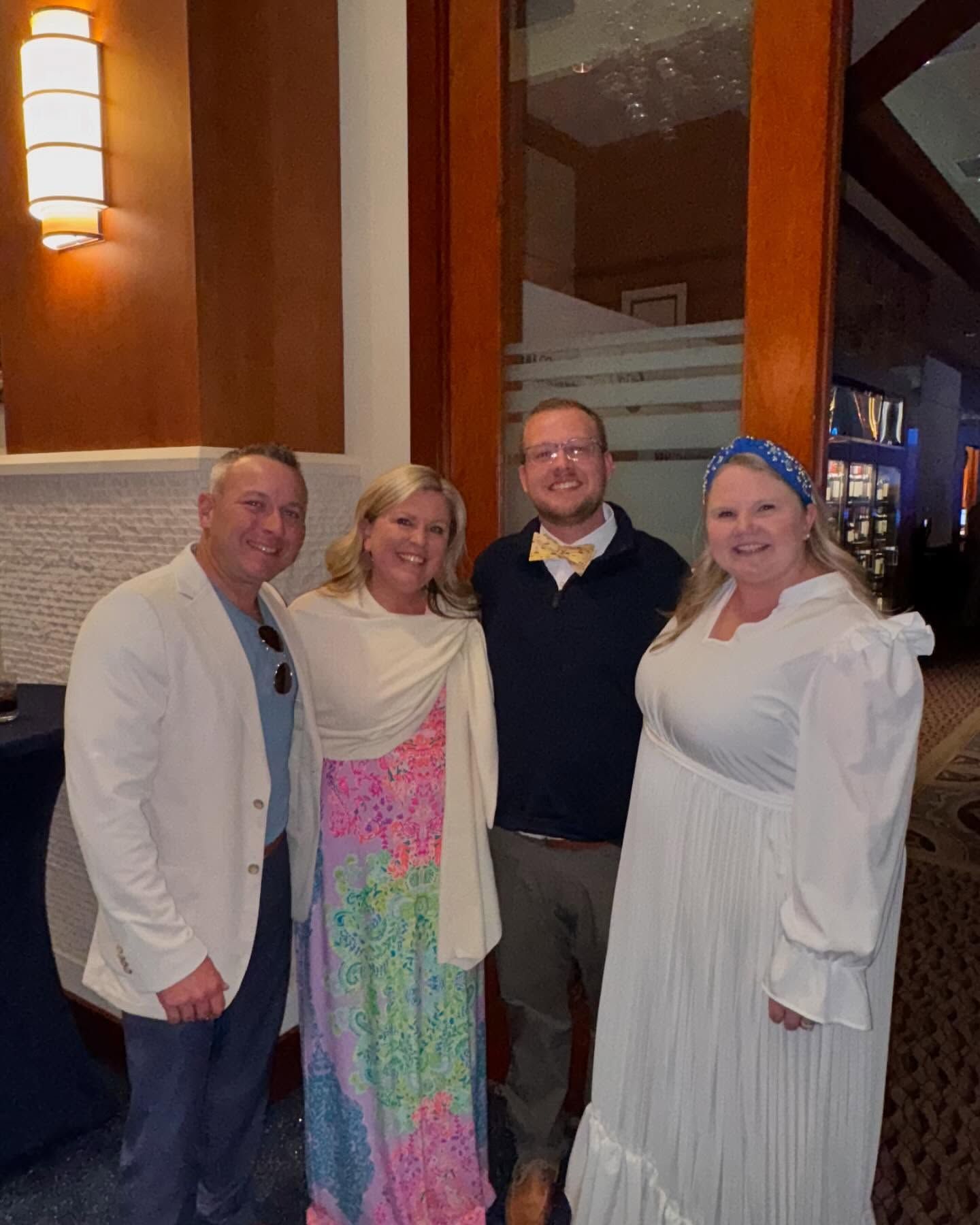 Four people posing for a photo. Indoors with wood and glass accents. One person in white blazer, one in colorful dress, one in sweater, one in white dress.