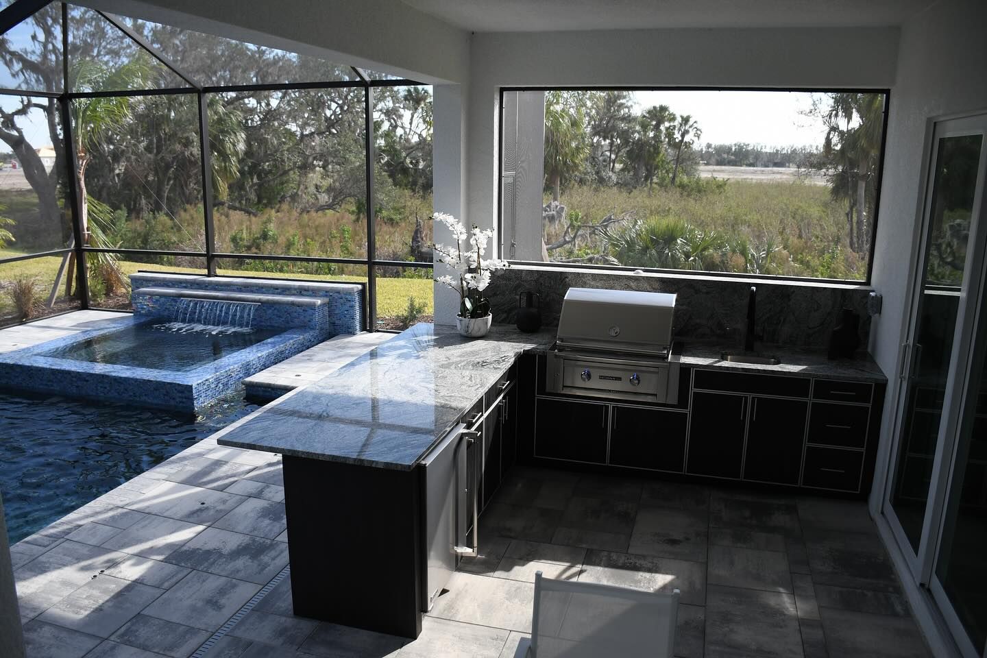 Outdoor kitchen with pool. Gray countertops, stainless steel grill, dark cabinets, and a pool with blue tile.