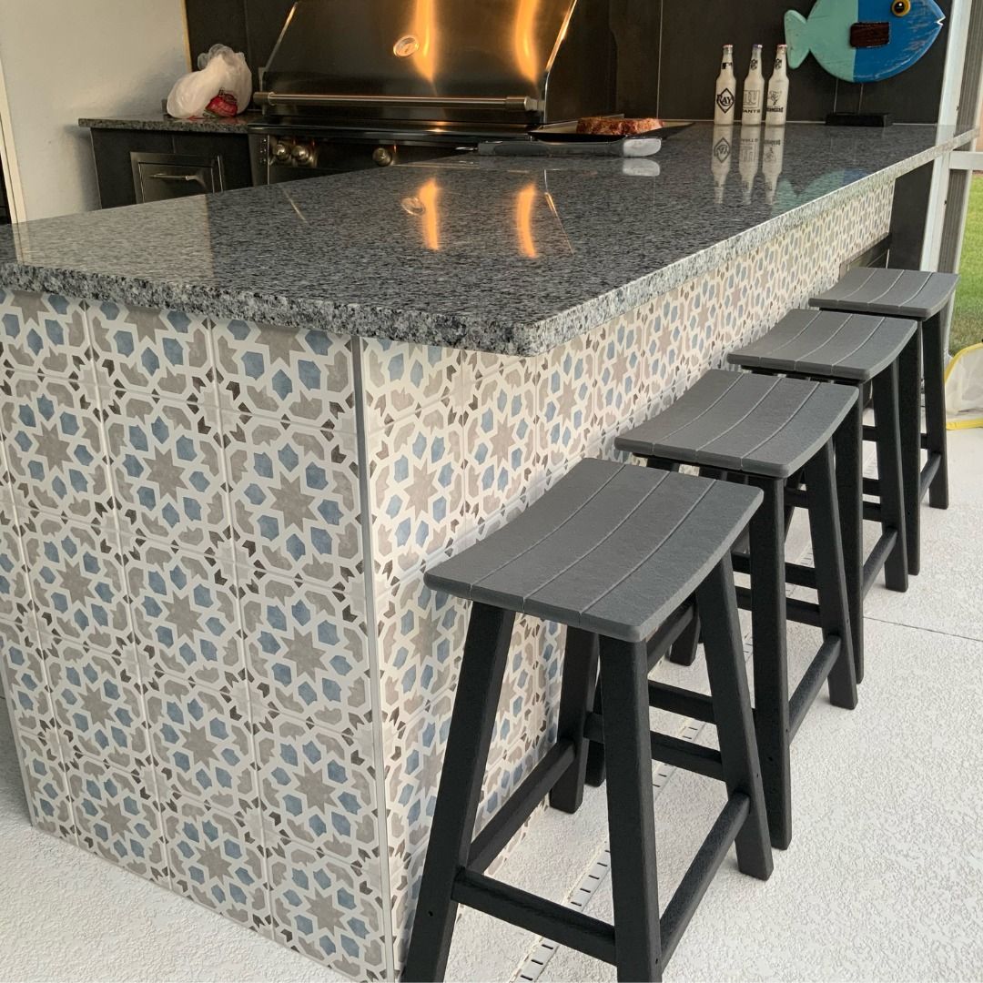Outdoor kitchen with granite countertop, patterned tile base, and four grey stools.