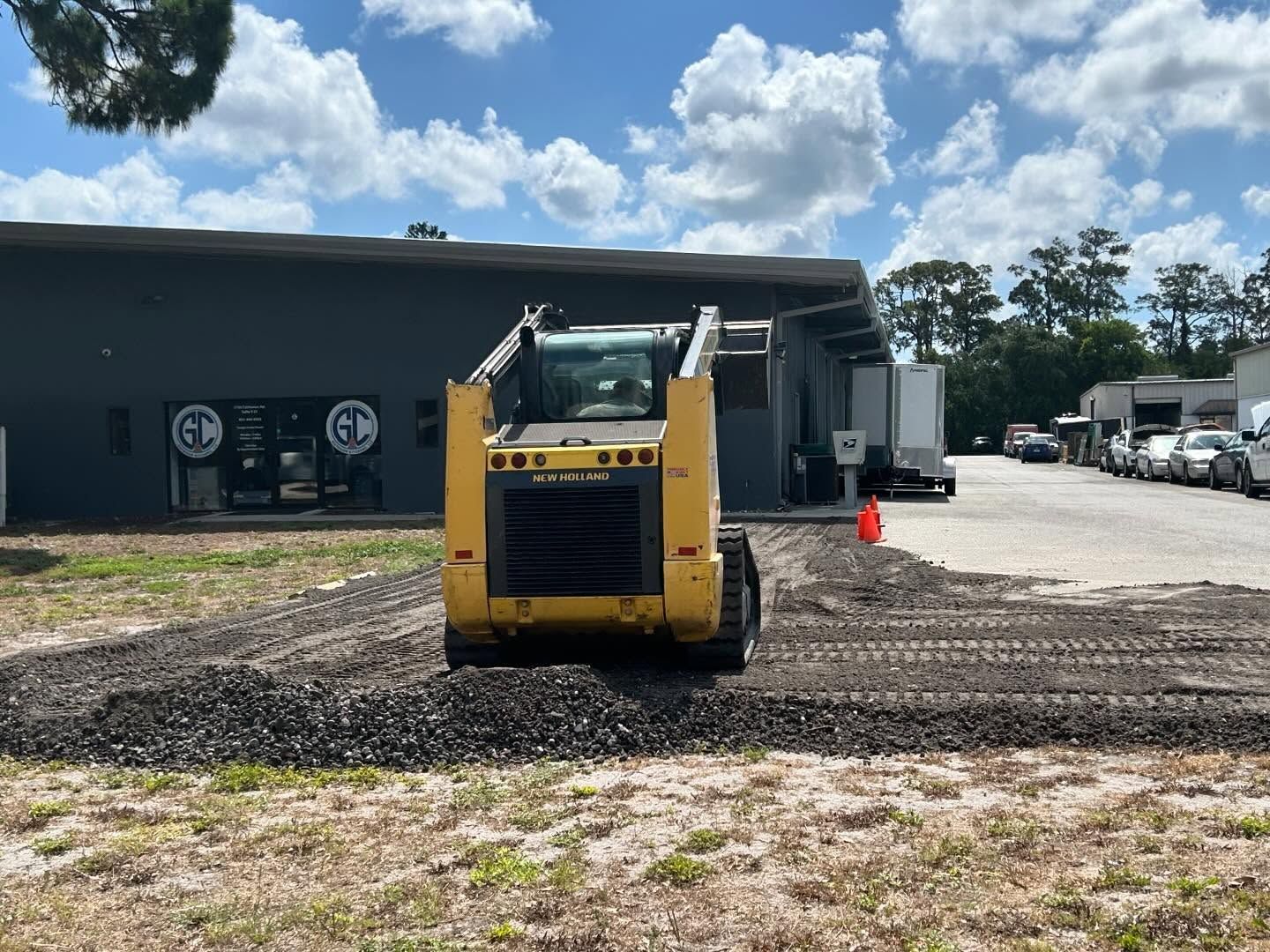 Yellow skid steer spreading gravel in front of a building with parked vehicles and blue sky.