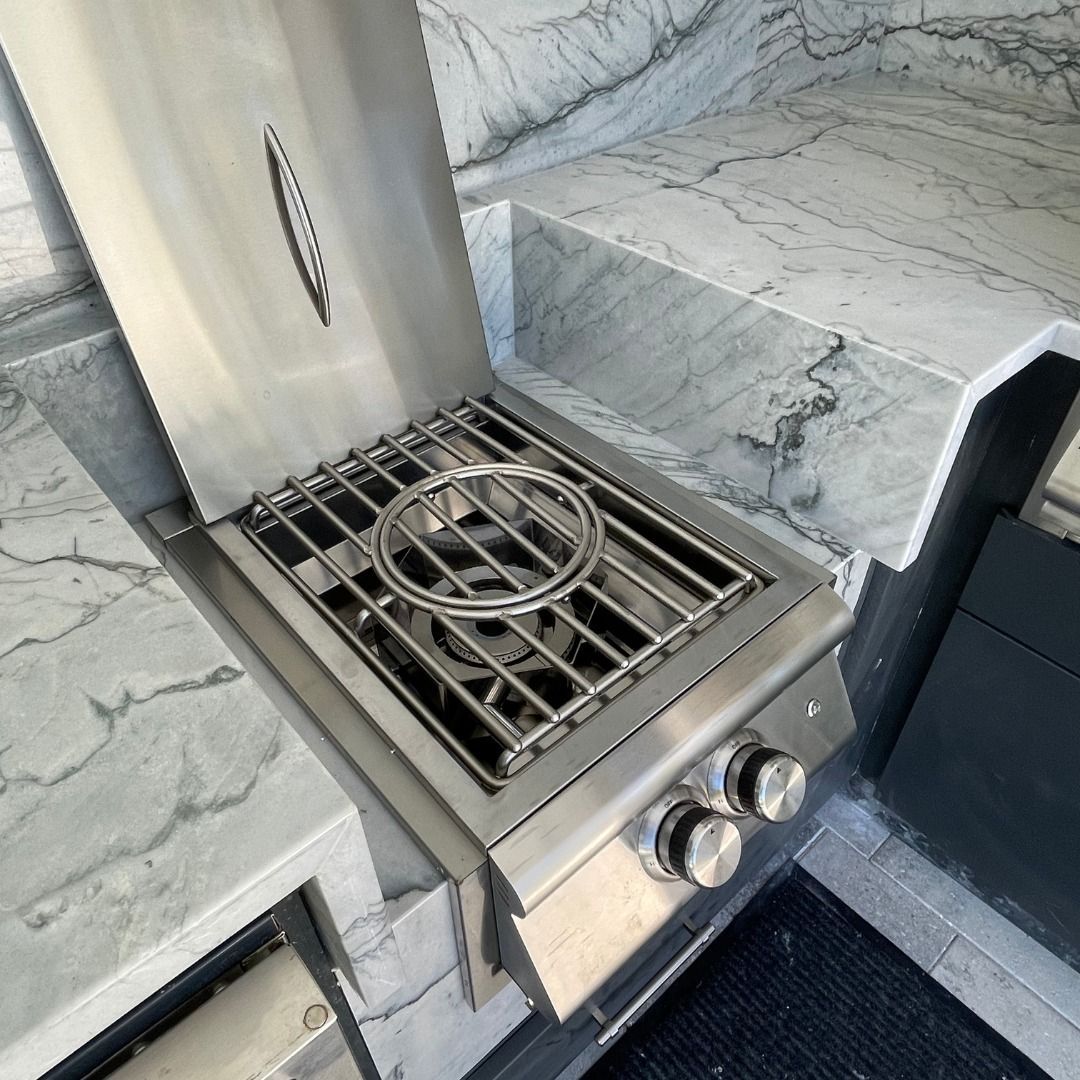 Stainless steel outdoor grill built into a marbled countertop. The lid is open, exposing the grill grates and burner knobs.