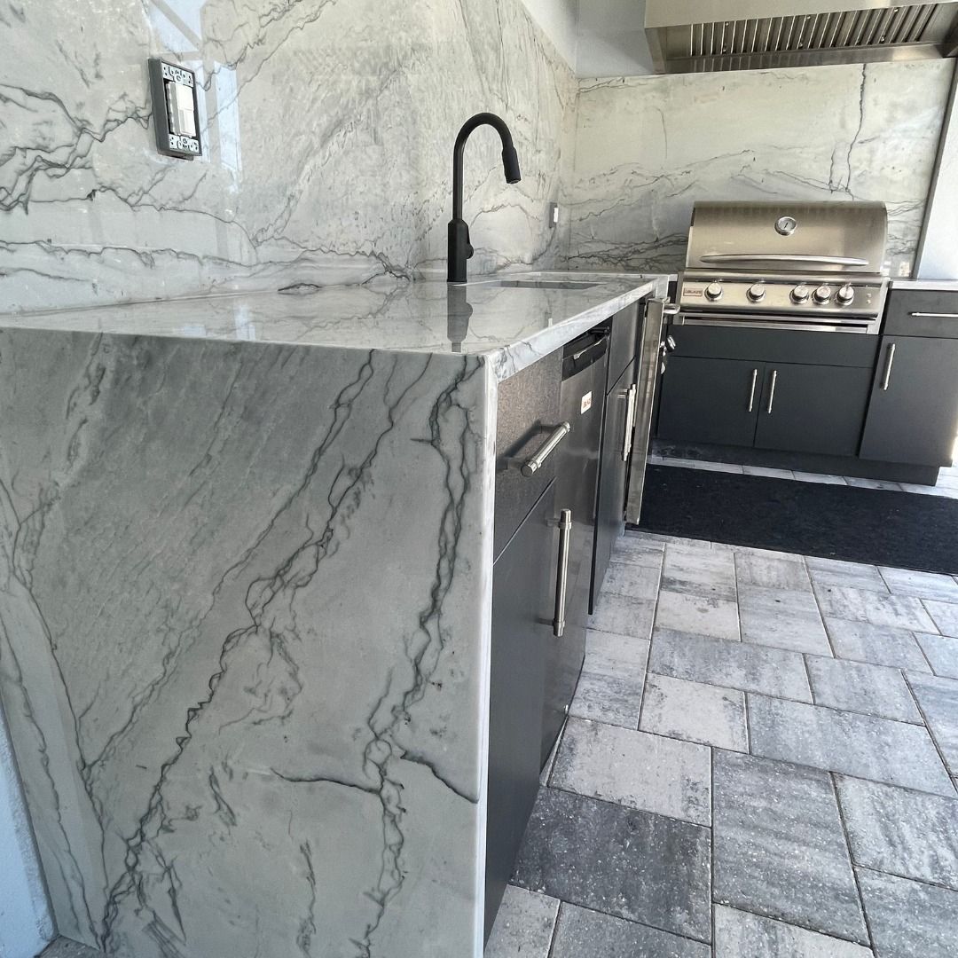 Outdoor kitchen with marble countertops, stainless steel grill, and gray cabinets.