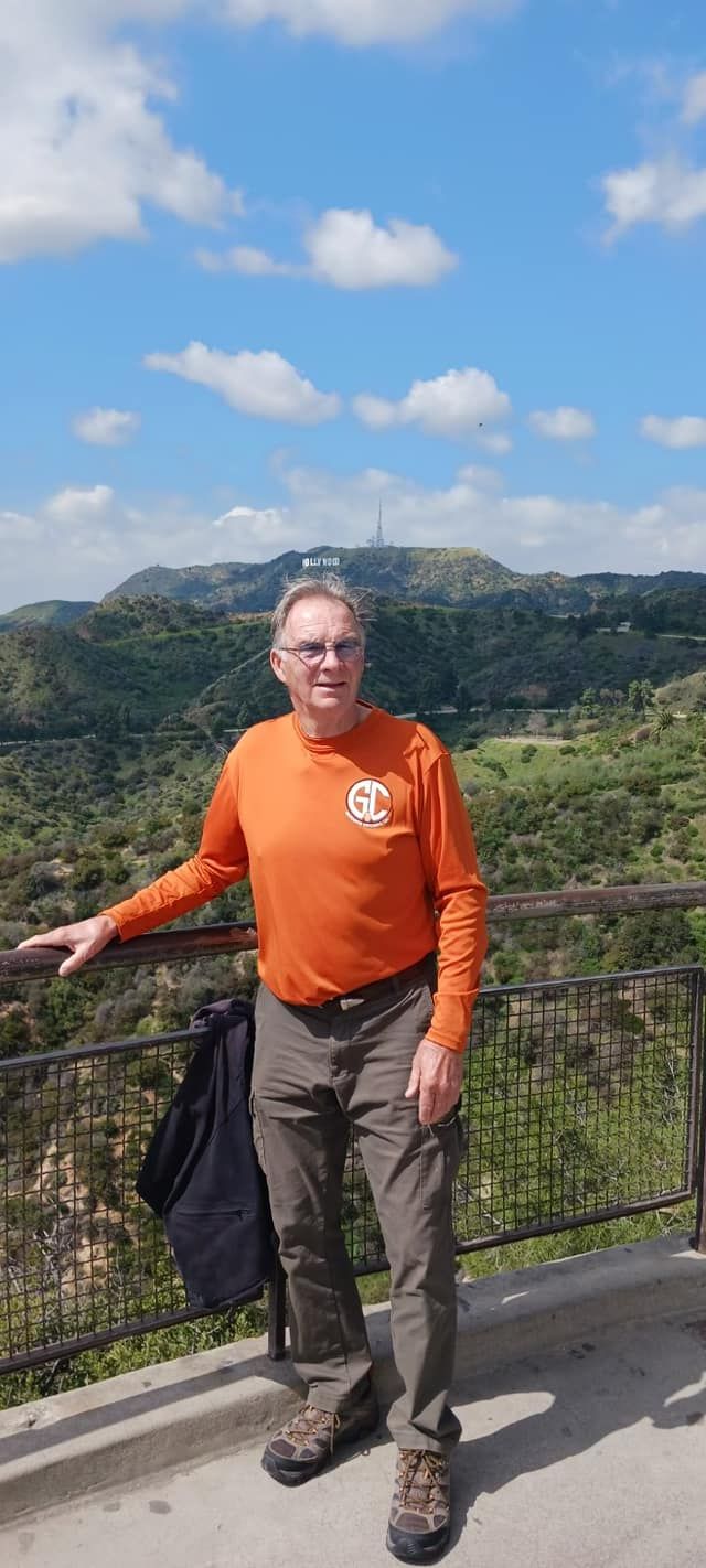 Man in orange shirt stands at a viewpoint, overlooking a valley under a cloudy blue sky.