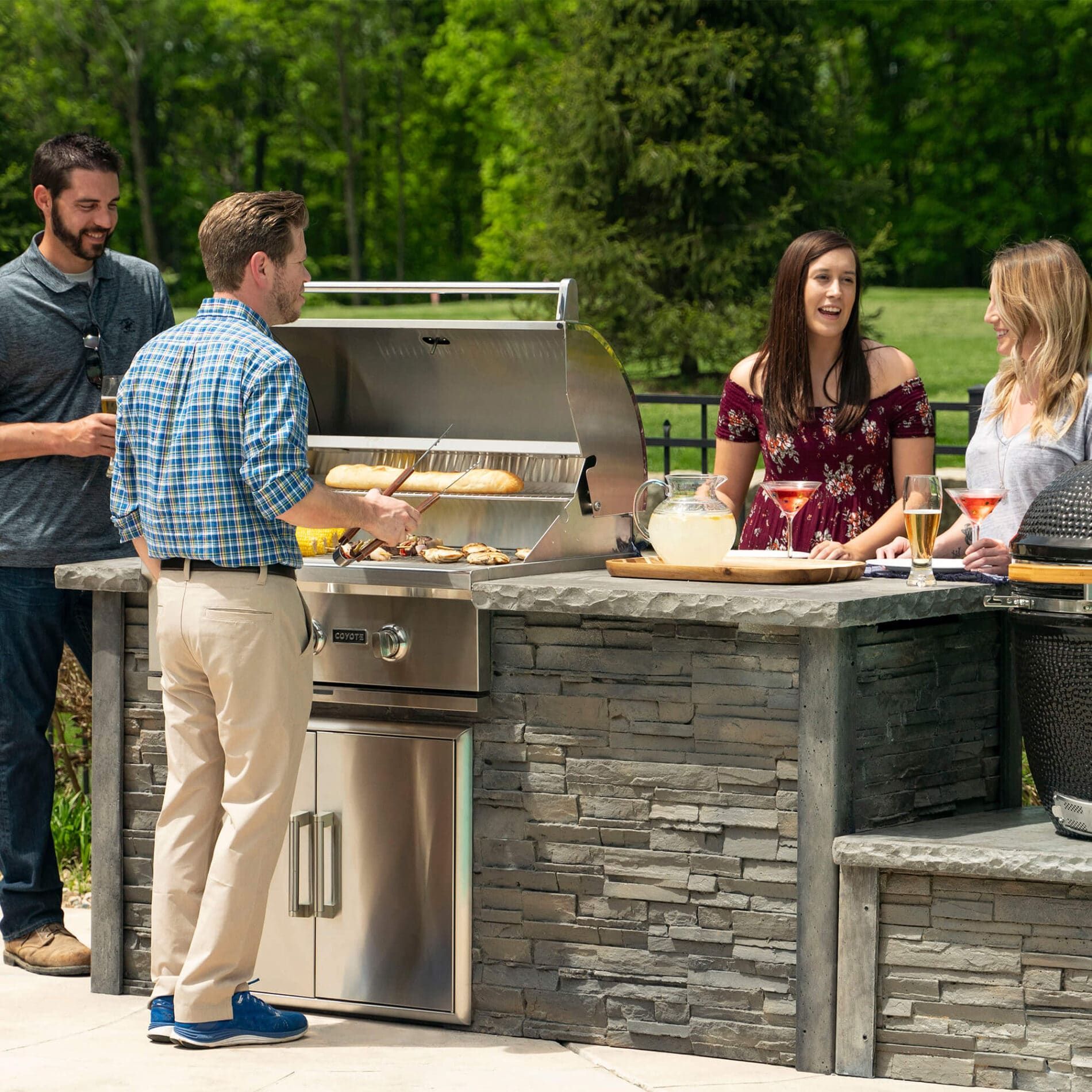 Four people at an outdoor kitchen with a grill. One person is cooking, others are smiling, talking, and holding drinks.