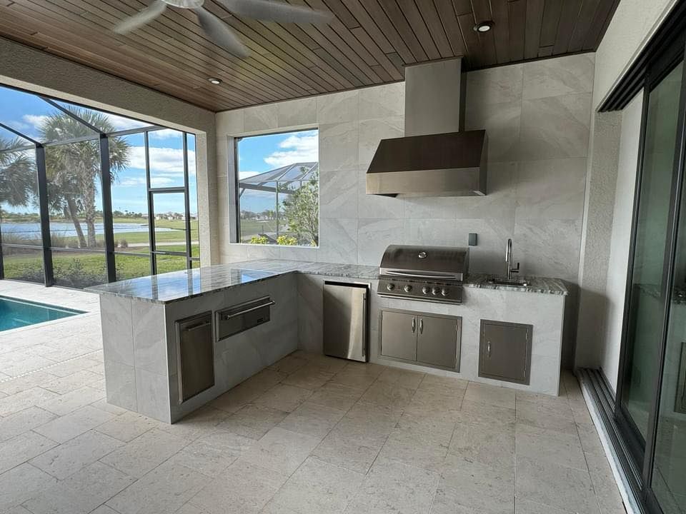 Outdoor kitchen with grill, hood, sink, and island; light gray tile, wood ceiling, view of water.