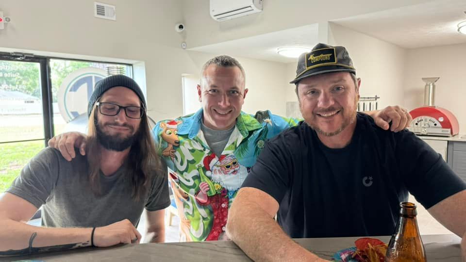 Three men smiling indoors, one with a Santa shirt, arms around the others.