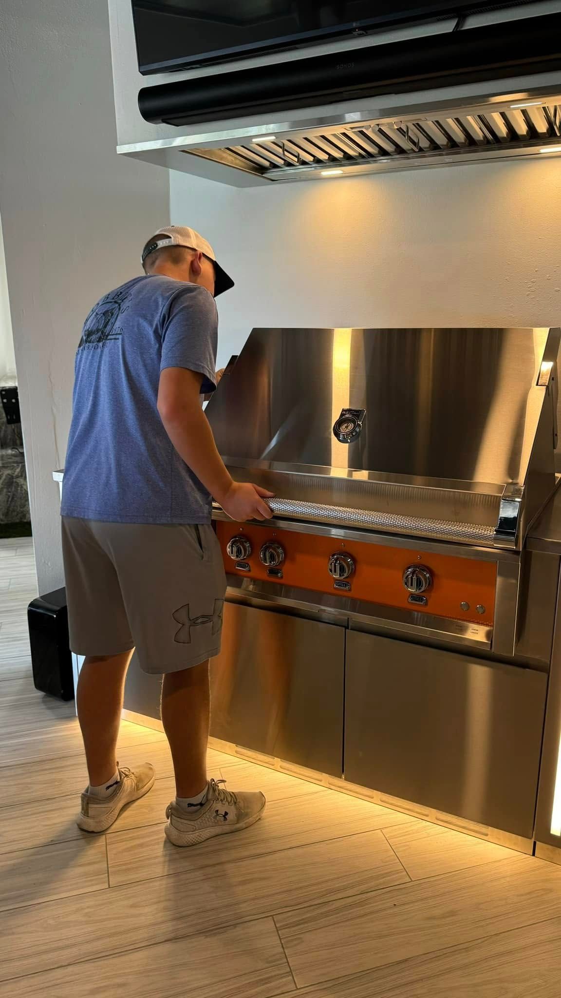 Man wearing a hat and shorts grilling food on an outdoor kitchen, looking at the camera.