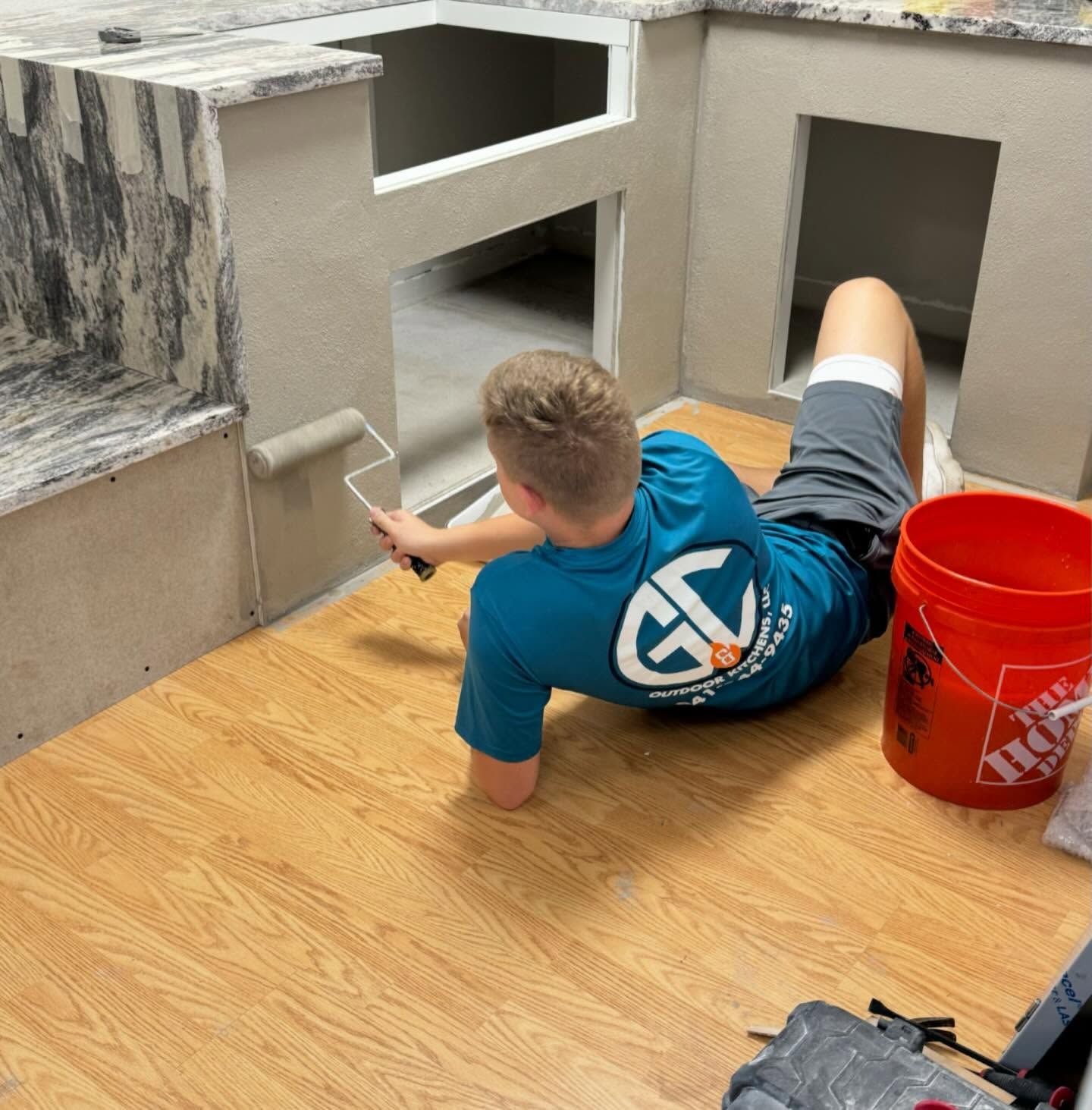 A person painting a wall while lying on a wood-look floor, with a bucket nearby.