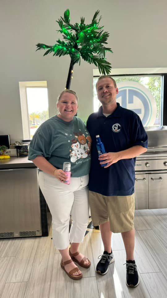 Two people smiling, holding drinks under a decorated palm tree. Inside with outdoor kitchen visible.