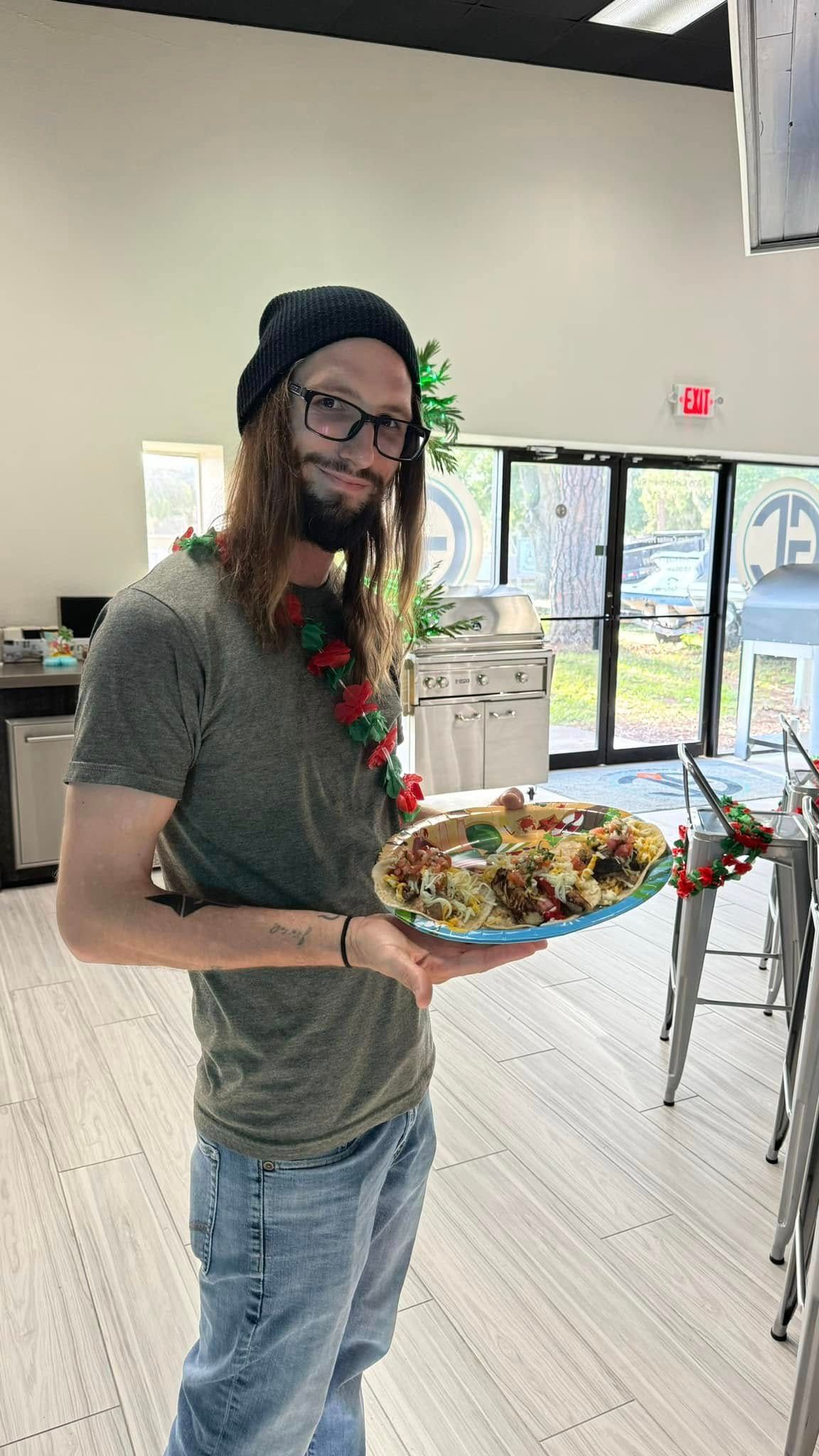Man holding a plate of food, wearing a beanie, glasses, and a garland. Indoors, smiling.