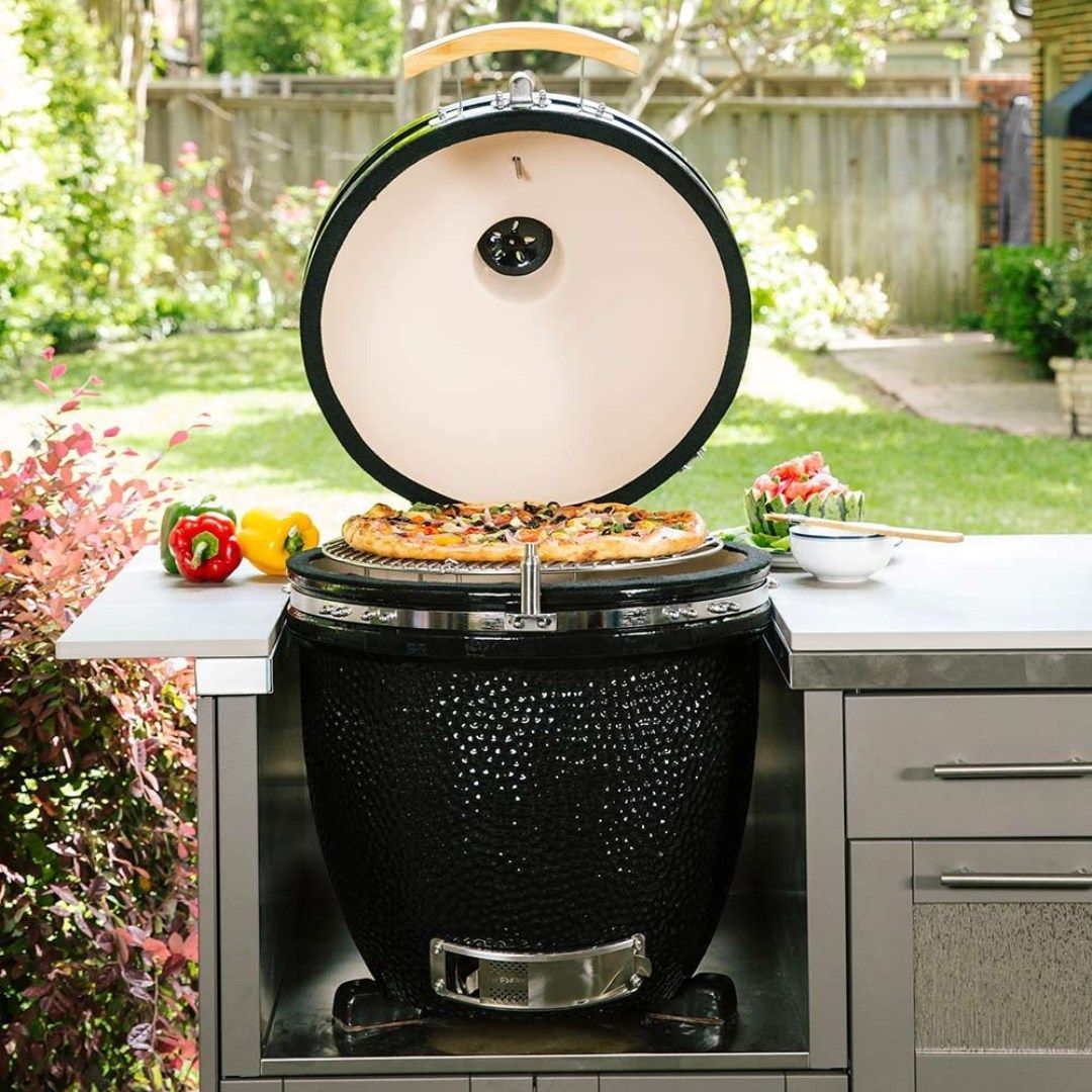 Black ceramic grill with open lid, cooking a pizza on a stainless steel countertop in a backyard setting.