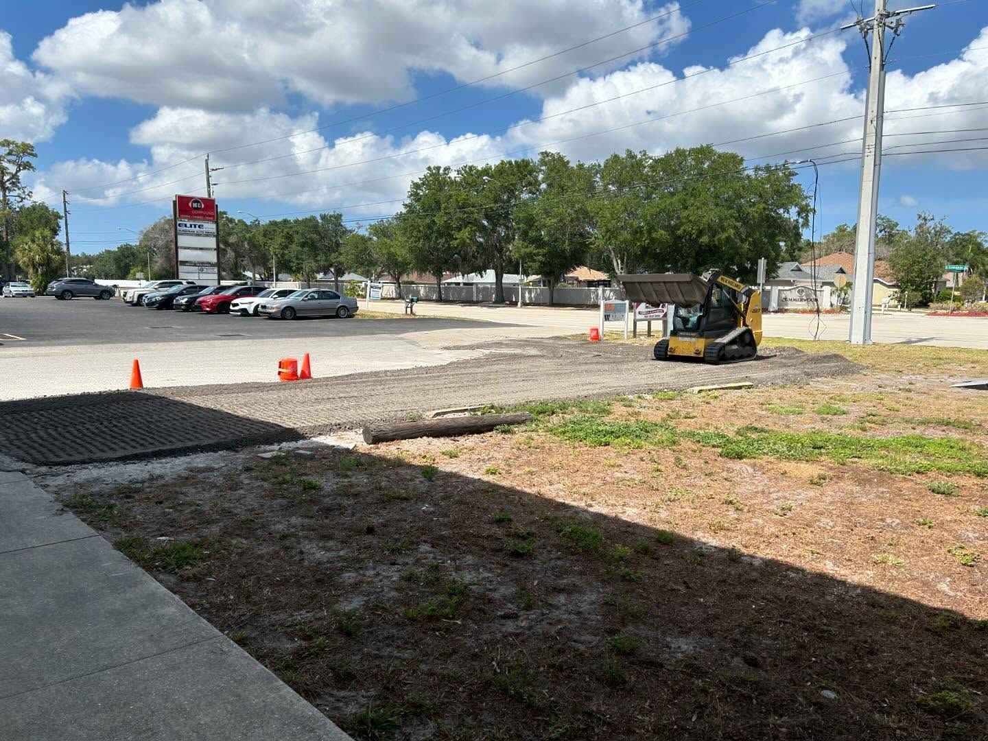 Construction site with small excavator, dirt, gravel, and cones. Cars in parking lot under cloudy sky.