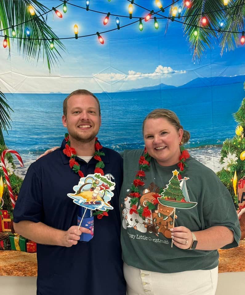 Two people smiling, posing in front of tropical Christmas backdrop, wearing leis, holding props.
