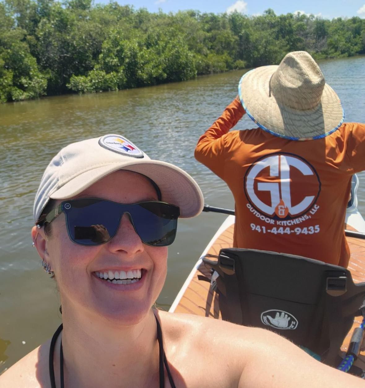 Woman smiling, wearing sunglasses and a hat, on a paddleboard with a person in an orange shirt on a sunny body of water.