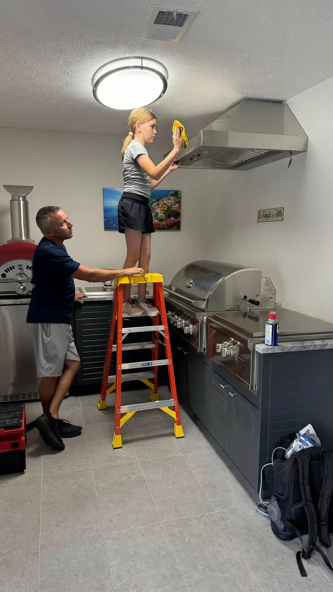 A person on a ladder cleans a range hood, assisted by another person in an outdoor kitchen.