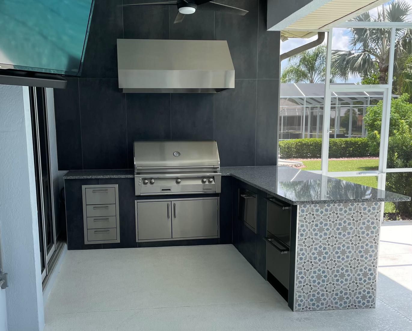 Outdoor kitchen with stainless steel grill, hood, and bar. Gray cabinets, countertop, and floor.