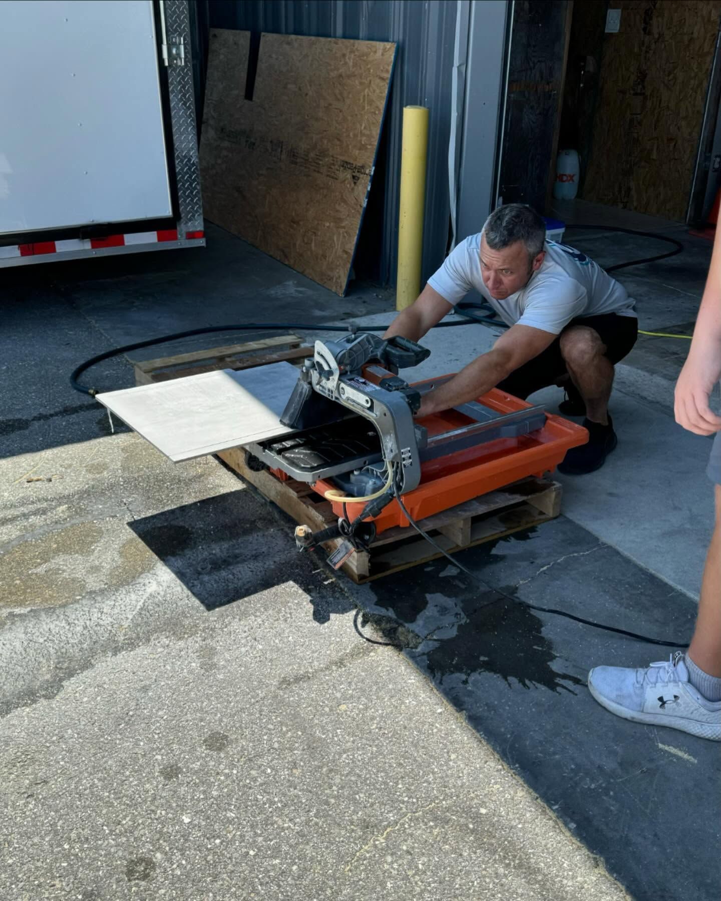 Man using a tile saw outdoors, cutting a tile. Water and sunlight visible.