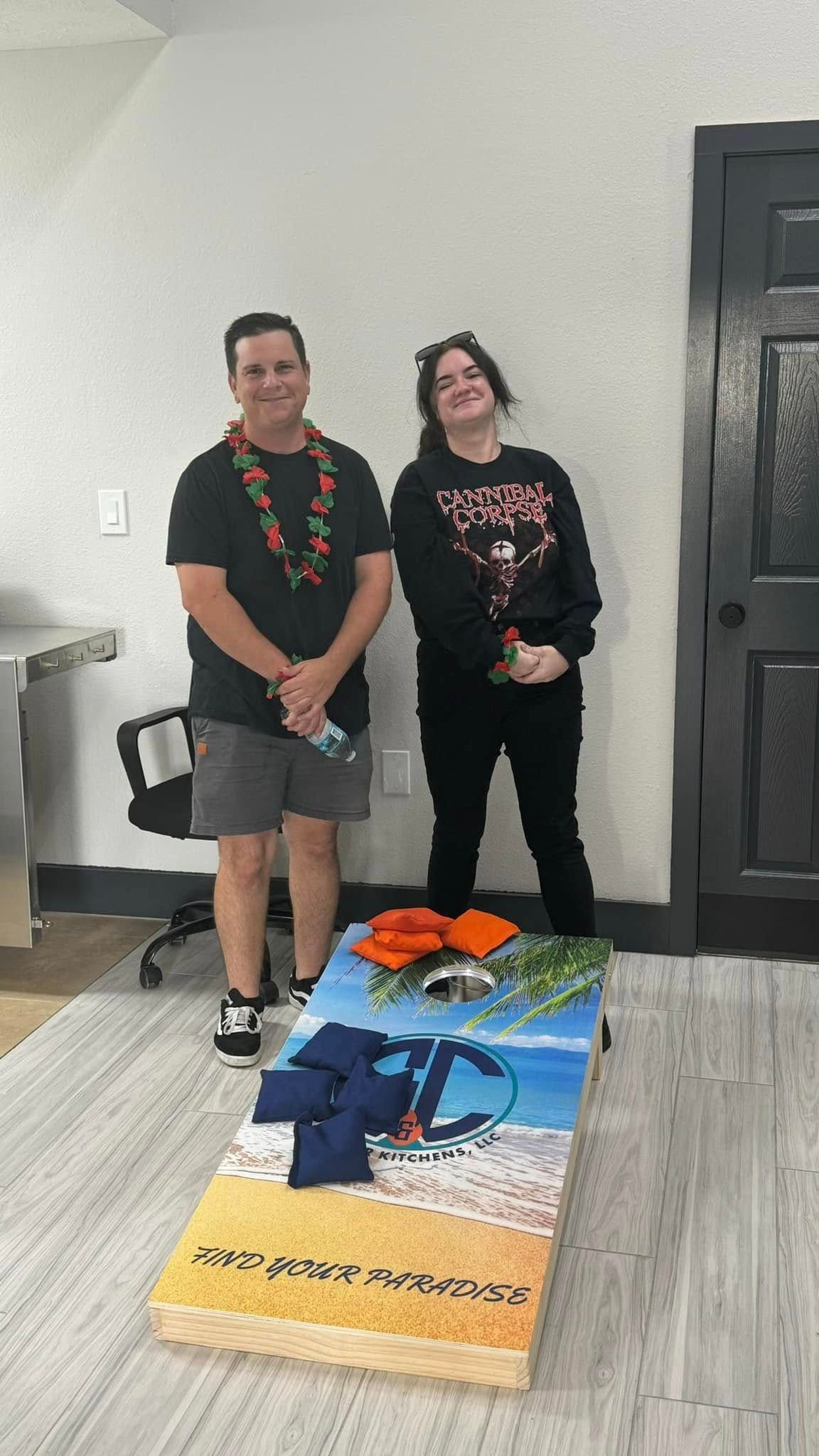Two people standing beside a cornhole board with a beach scene design; one person has a lei.