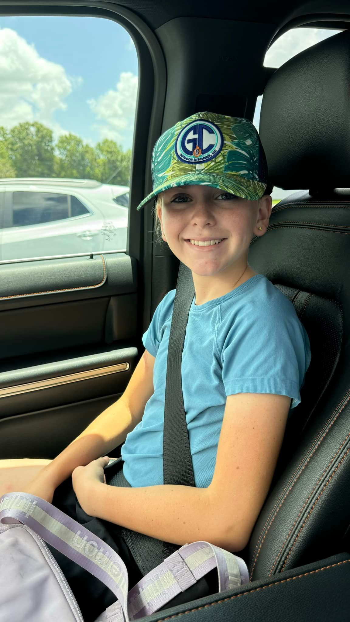 Girl wearing a green patterned hat and blue shirt smiles, seated in a car, seatbelt on.