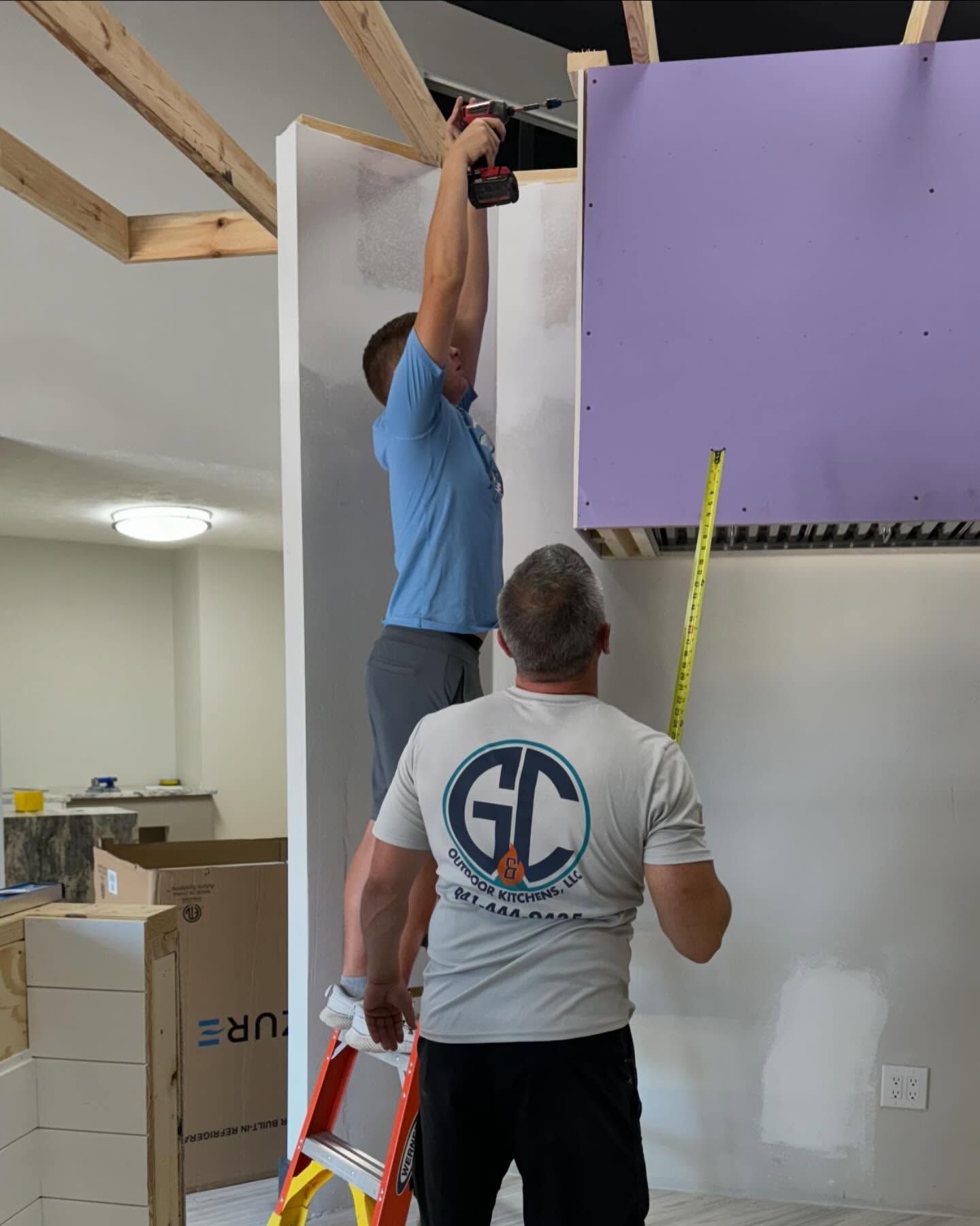 Two people installing drywall; one on a ladder using a drill, the other measuring.