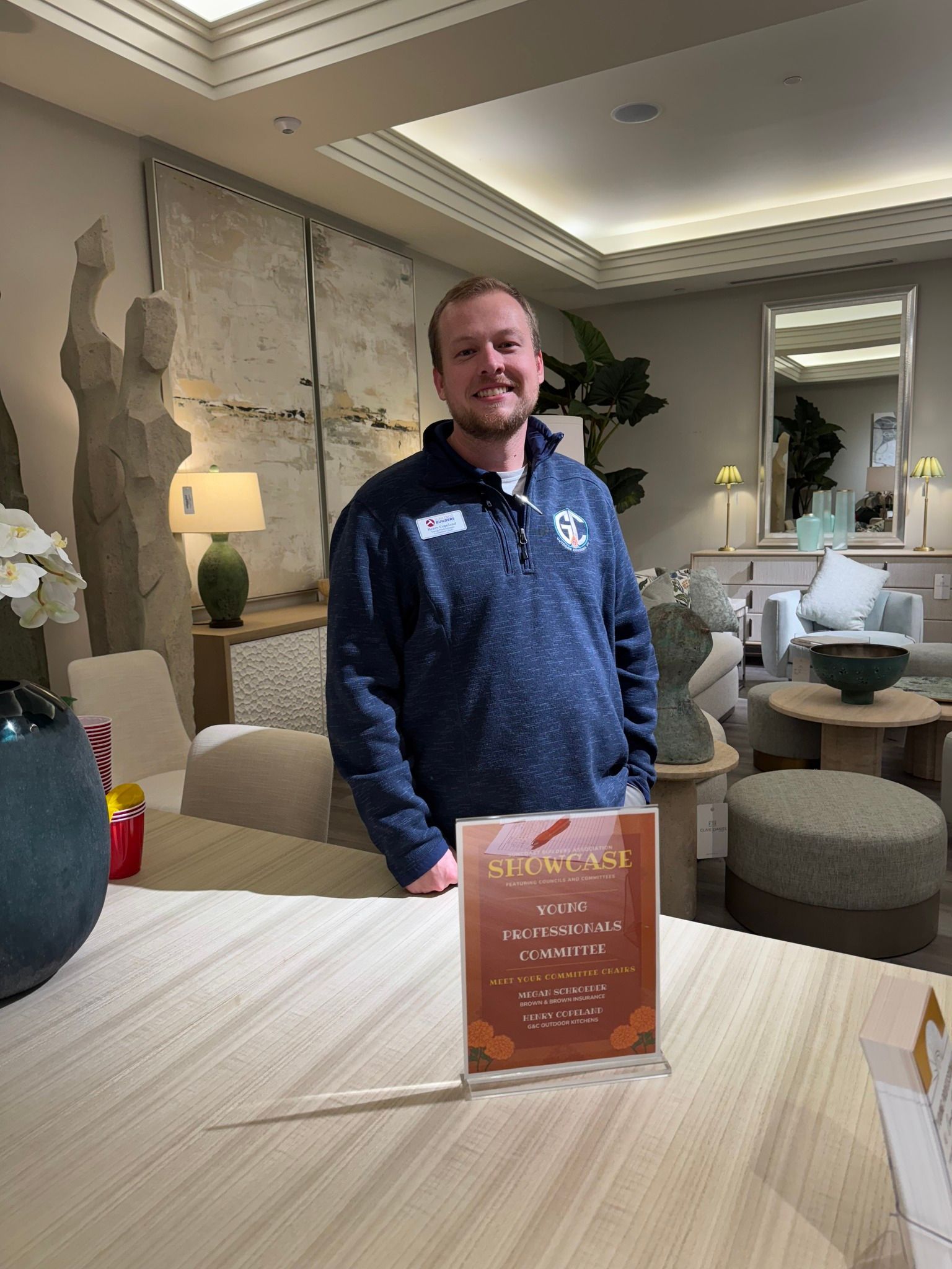 Man in blue sweater stands behind a table in a furniture store, near a sign.