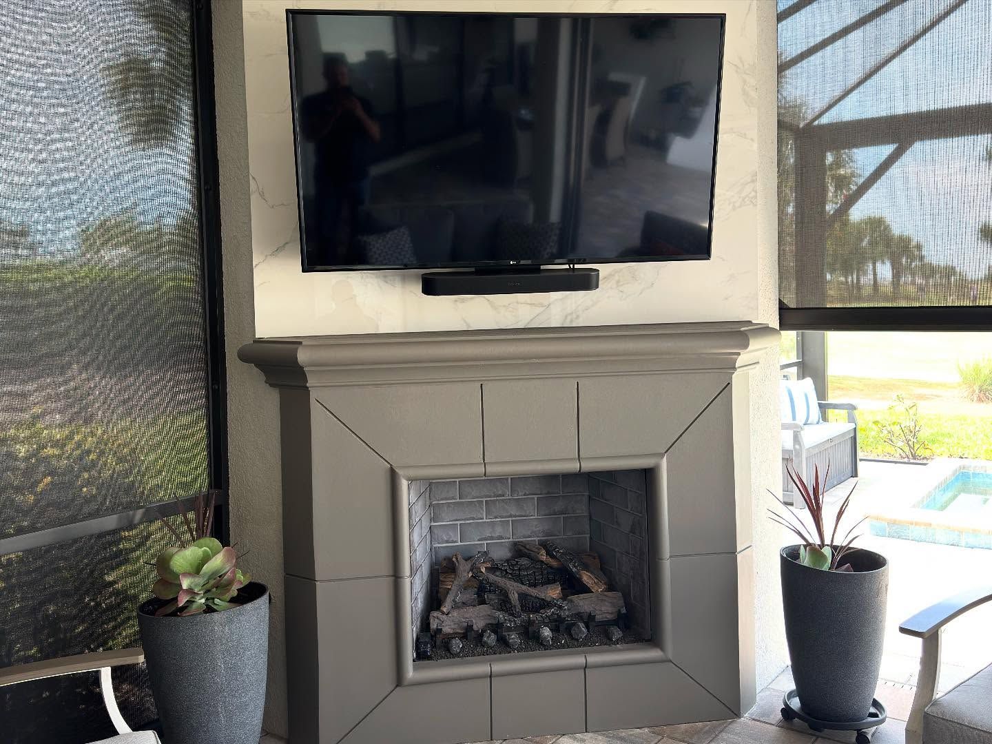 Outdoor fireplace with mounted TV, gray surround, and potted plants.