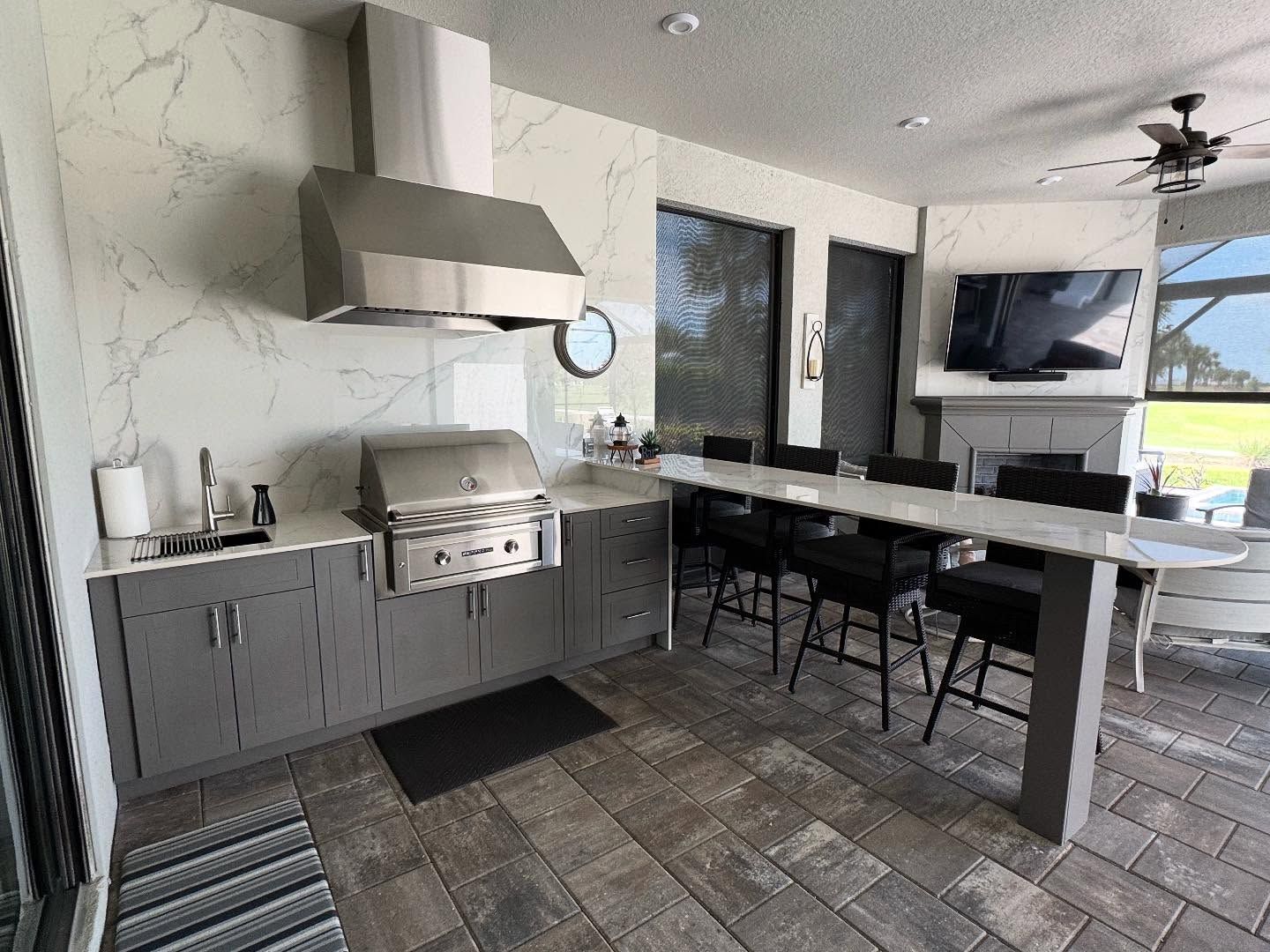Outdoor kitchen with grill, hood, countertop, bar stools, and TV, gray cabinets and marble-look backsplash.
