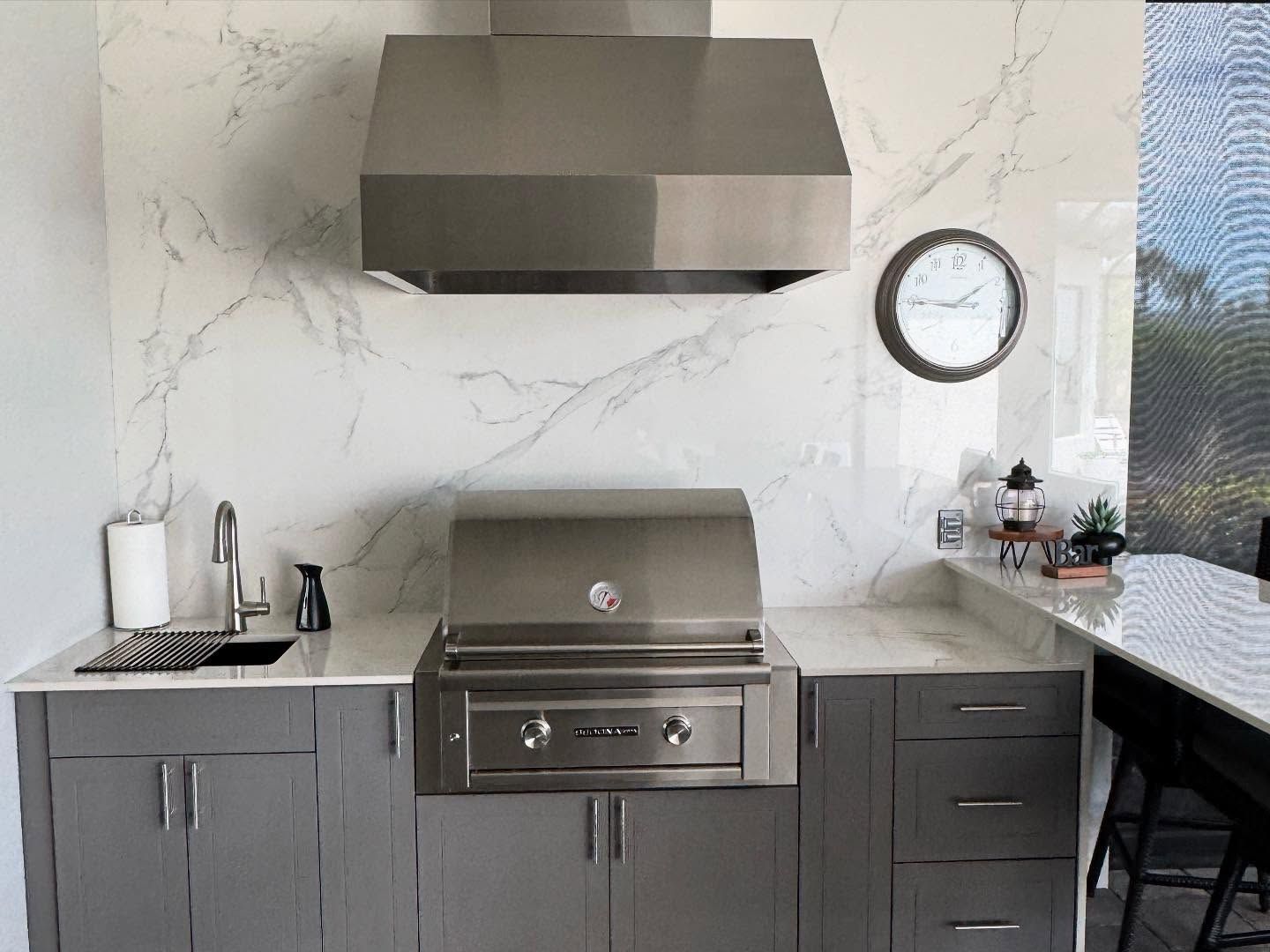 Outdoor kitchen with stainless steel grill, hood, sink, gray cabinets, and marble-look backsplash.