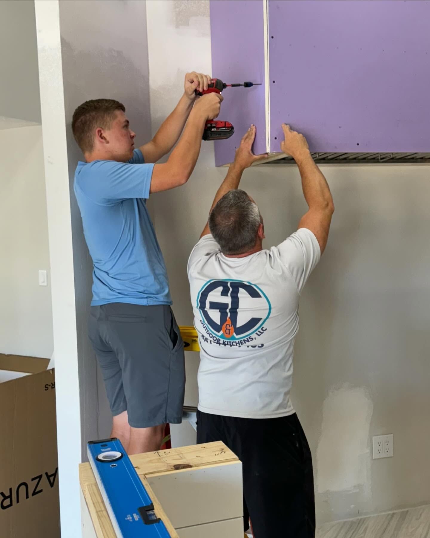 Two people installing purple drywall with a drill and screw, indoors.