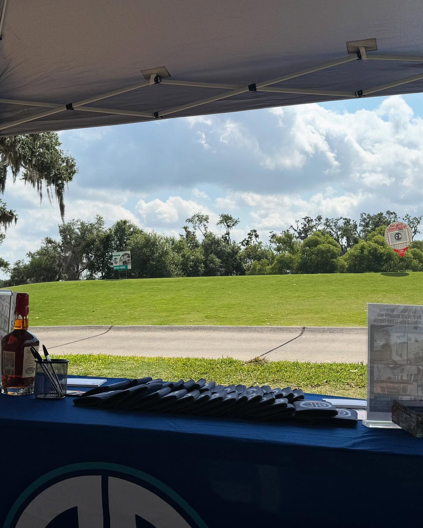 Outdoor table with bottles, glasses, and a stack of black objects, with a green field and trees in the background under a cloudy sky.